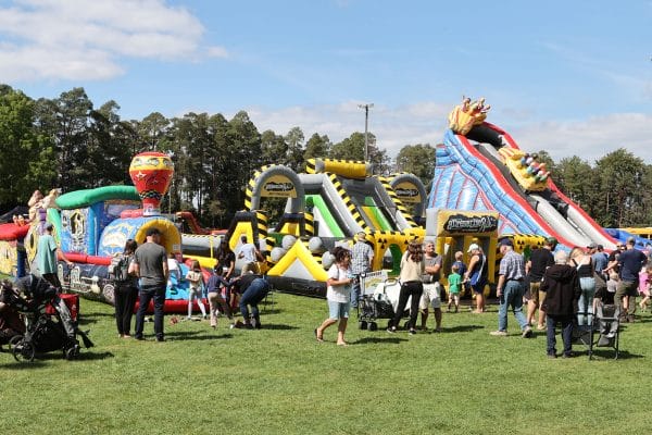 Large colorful inflatable play park with multiple bounce houses, obstacle courses, and slides set up on green grass at the Caledon Fair, with families and children enjoying the attractions under a blue sky with trees in the background.