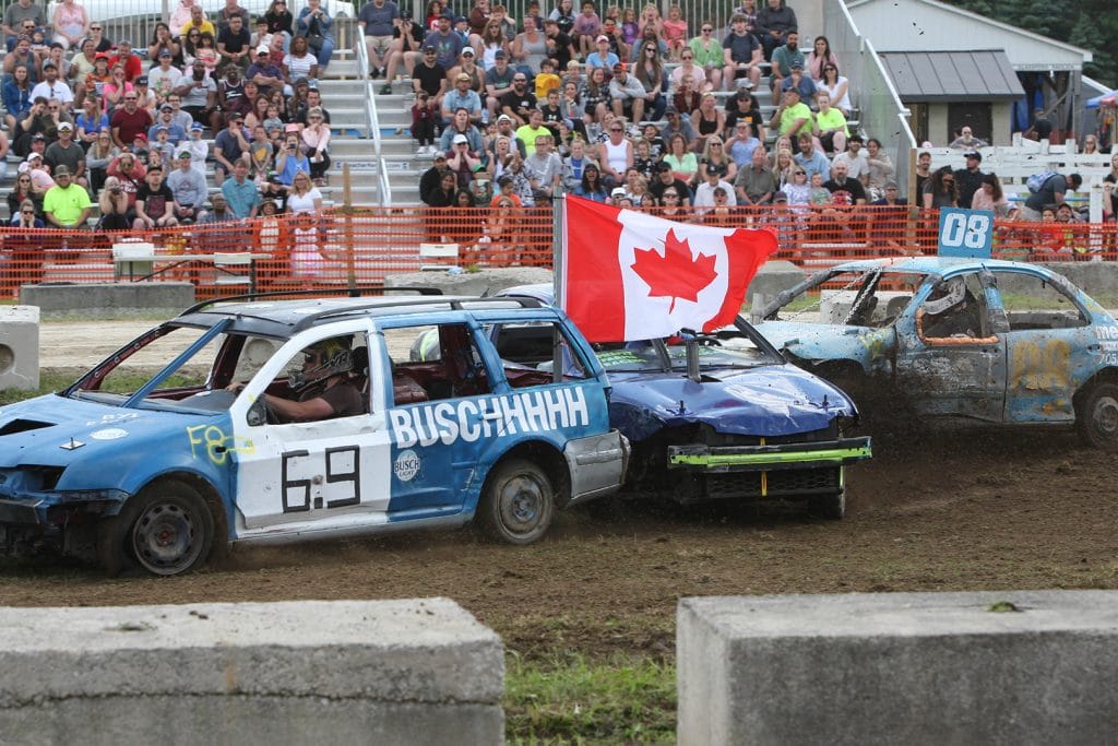 Demolition derby car number 69 with "BUSCHHHHHH" written on the side proudly displays a large Canadian flag while competing at the Caledon Fair, with packed grandstands of spectators in the background watching the exciting motorsport event.