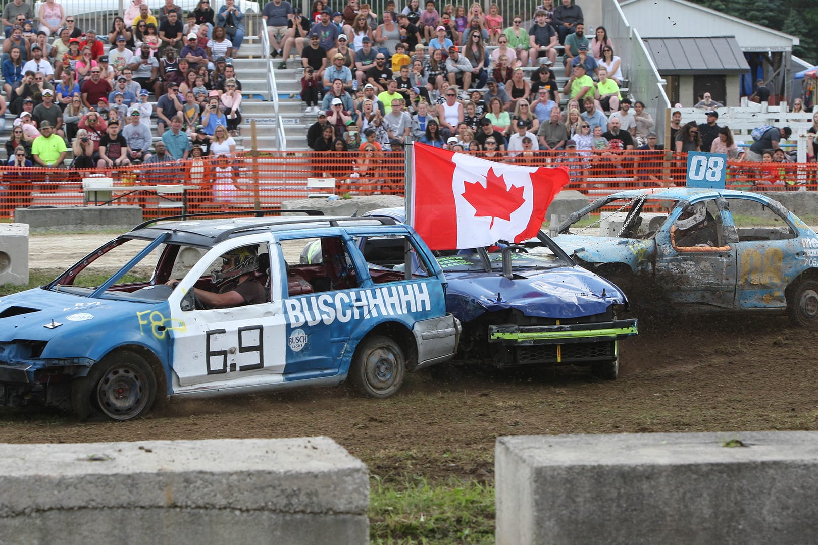 Demolition derby car number 69 with "BUSCHHHHHH" written on the side proudly displays a large Canadian flag while competing at the Caledon Fair, with packed grandstands of spectators in the background watching the exciting motorsport event.
